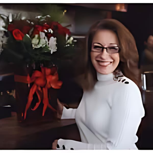 Smiling woman in glasses and a white turtleneck next to a bouquet of red and white flowers.