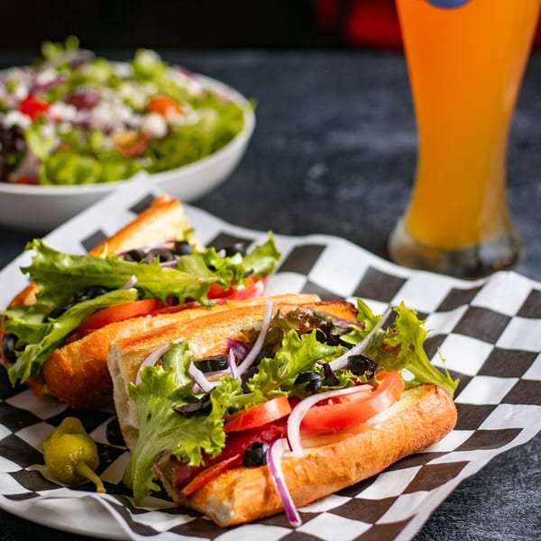 Two Italian subs with lettuce, tomatoes, onions, and olives, served with a side salad and beer.