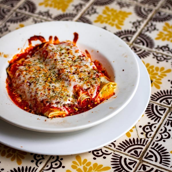 Baked manicotti with melted cheese, red sauce, and herbs in a white bowl on a patterned tile table.