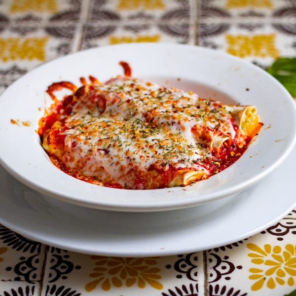 Baked manicotti or cannelloni with melted cheese, red sauce, and herbs in a white bowl.