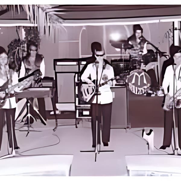 A band performing on stage with a drummer, guitarist, and keyboardist.
